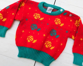 Puede incluir: Jersey de punto rojo con ribete verde y estampado floral amarillo y verde. El jersey tiene una etiqueta de talla que dice "M".