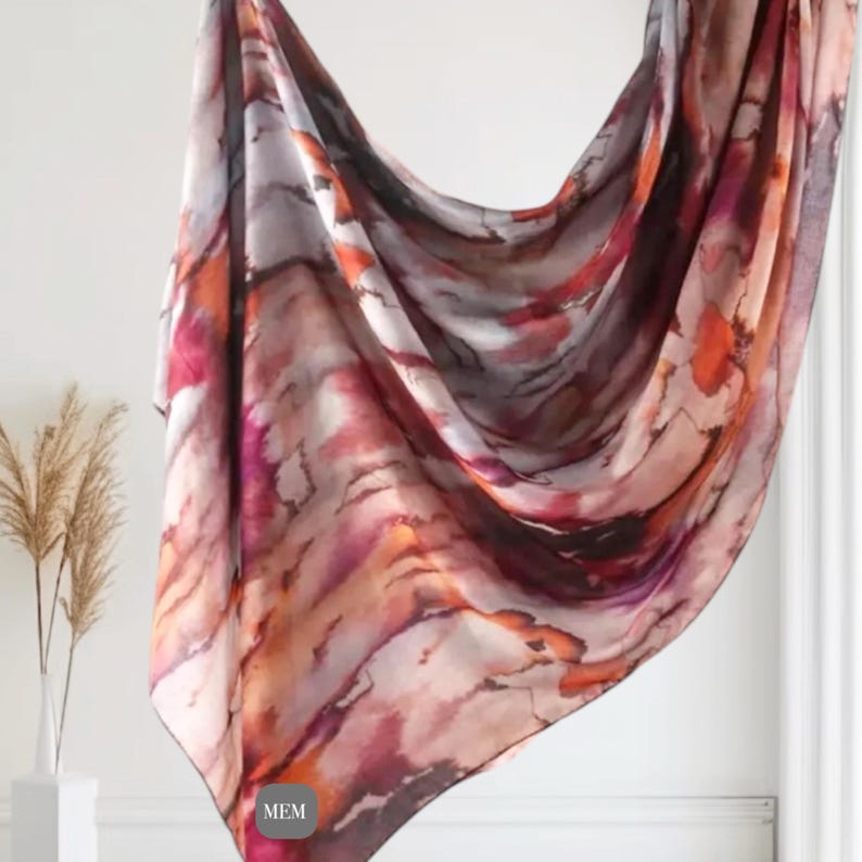 May include: A large, rectangular scarf with a watercolor-like print in shades of orange, pink, and brown. The scarf is made of a soft, flowing fabric and has a subtle sheen.