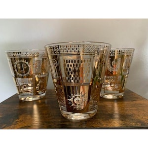 May include: Three clear drinking glasses with gold geometric and sunburst designs. The glasses are on a wooden surface. The glasses have a vintage aesthetic.