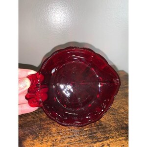 May include: A deep red, translucent glass dish with a scalloped edge and a small, integrated handle on one side. The circular bowl features a smooth, reflective surface and a molded design that catches light, creating varied shades of crimson.