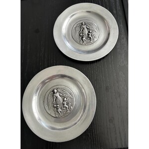 May include: Two pewter plates with a raised relief design. Each plate features a central medallion with an angel figure and several smaller figures. The plates have a simple, circular design and a slightly raised rim.