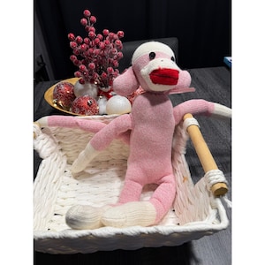 May include: A pink sock monkey doll with white accents, red lips, and black button eyes. The monkey is in a white woven basket with a wooden handle. Decorative red berries and ornaments are in the background.