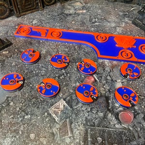 Può includere: Un tabellone da gioco in plastica blu e arancione con un design a forma di teschio e ossa incrociate. Il tabellone ha un bordo rialzato e un'apertura al centro. Ci sono otto gettoni in plastica blu e arancione più piccoli con design a forma di teschio e ossa incrociate.