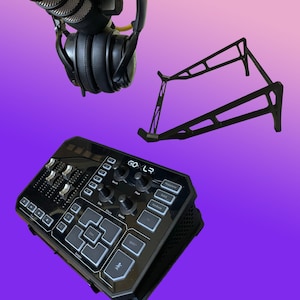 Może przedstawiać: Czarny mikser audio GoXLR Mini z różnymi przyciskami i pokrętłami. Mikser ma czarny stojak na słuchawki w tle.