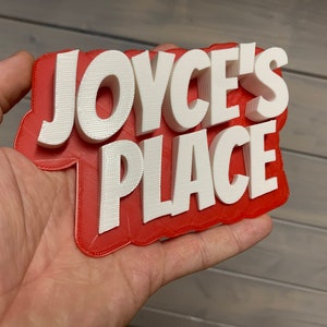 May include: A 3D-printed sign with the text "JOYCE'S PLACE" in white, raised letters against a red background. The sign is held in a hand, showcasing the layered design and texture of the plastic.