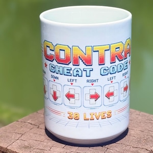 May include: A white ceramic mug featuring the text CONTRA CHEAT CODE in pixelated font. Below the text, a sequence of directional arrows and buttons is displayed, followed by the text 30 LIVES in a pixelated orange and yellow gradient font.