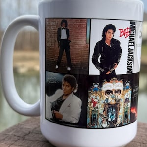 May include: White ceramic mug featuring multiple images of Michael Jackson, including album art and iconic poses. The mug has a large handle and is displayed on a wooden surface.