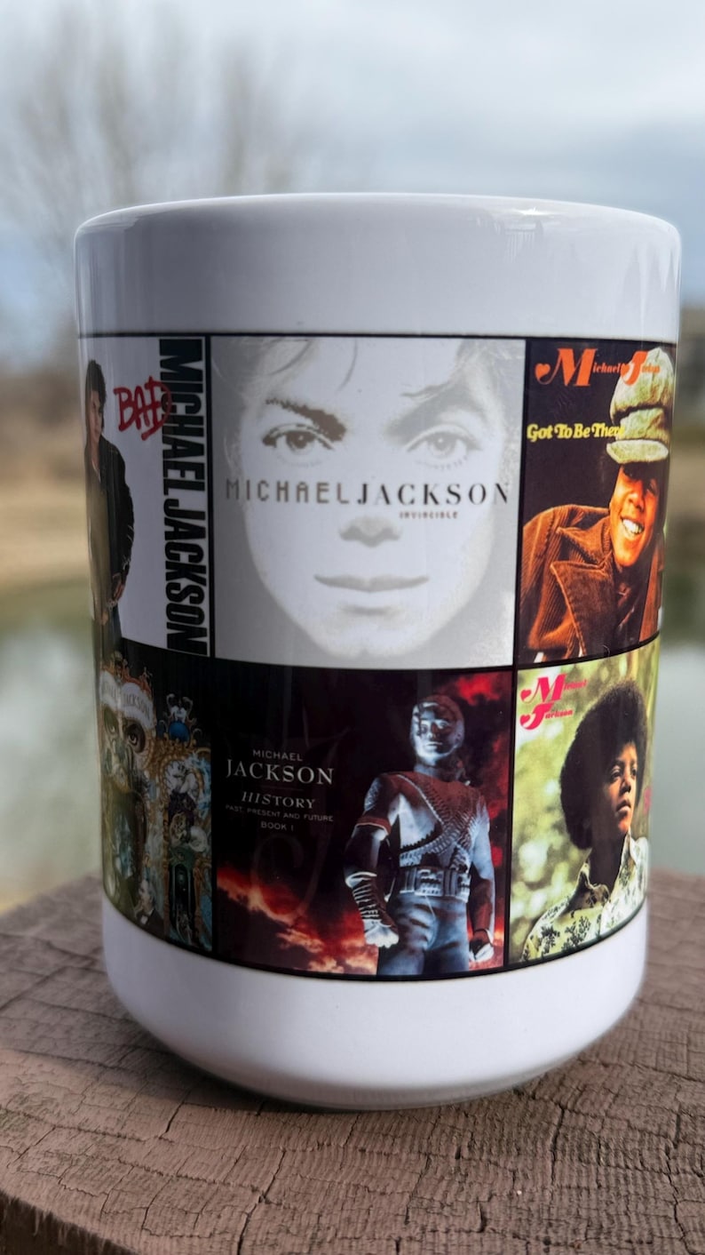 May include: White ceramic mug featuring a collage of Michael Jackson album covers and images. The mug includes album titles like "Bad" and "Invincible". The mug is cylindrical and sits on a wooden surface.