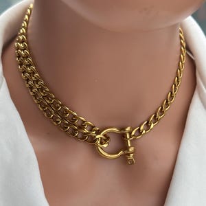 Peut inclure: Un collier en chaîne doré avec un design à double rang. Le collier présente une grosse chaîne gourmette et une fermeture à fermoir unique. Le bijou est présenté sur un fond blanc.