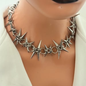 Puede incluir: Un collar gargantilla plateado con púas en forma de estrella. El collar está hecho de anillos de metal unidos y se usa alrededor del cuello. Las púas están espaciadas uniformemente y apuntan hacia afuera. El collar se usa sobre una prenda blanca.