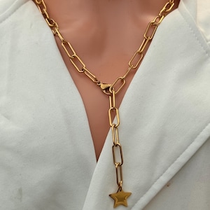 Può includere: Collana color oro con una catena a maglie allungate. Una catena verticale con un ciondolo a forma di stella pende dal centro. La collana è indossata sopra un capo bianco.