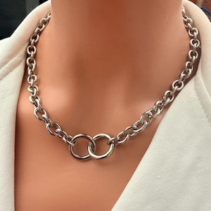 Op de afbeelding: Een zilverkleurige ketting met een reeks dikke, afgeronde schakels. Het midden van de ketting is voorzien van twee in elkaar grijpende cirkelvormige ringen die als middelpunt dienen en de twee zijden van de zware, gepolijste metalen ketting verbinden.