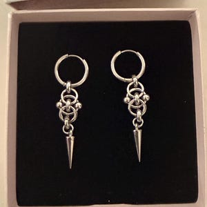 May include: A pair of silver-toned hoop earrings with a chainmail design and a conical spike pendant. The earrings are displayed in a black box with a pink interior.