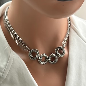May include: A silver-toned necklace featuring a chain design with a series of interlocking rings at the center. The necklace is displayed on a mannequin wearing a white shirt. The necklace has a modern, geometric aesthetic.