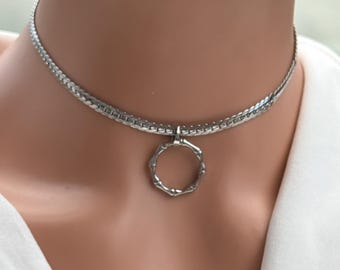 May include: A silver-toned choker necklace with a geometric pendant. The necklace features a woven chain design and a small, open, octagonal pendant. The jewelry is displayed on a person wearing a white top.