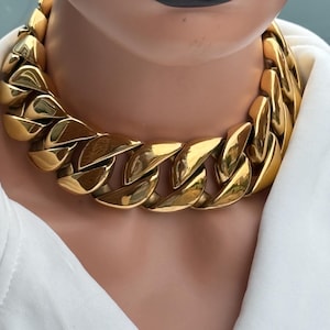 Puede incluir: Un collar de cadena dorado y grueso. El collar presenta eslabones grandes e interconectados y se usa sobre una camisa blanca. El modelo tiene lápiz labial negro. Una pieza de joyería audaz.