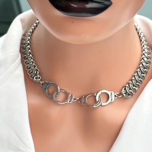 May include: A silver chain necklace with a handcuff pendant. The necklace has a thick, woven chain design and a central handcuff clasp. The image shows the necklace on a mannequin, with a white shirt and black lipstick.
