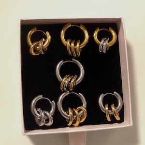 Puede incluir: Un conjunto de pendientes de aro en una caja de regalo. Los pendientes son dorados y plateados, con un diseño de triple anillo. La caja es rosa claro con el interior negro.