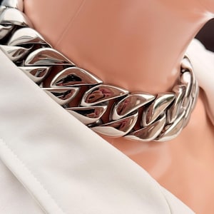 Puede incluir: Un collar de cadena plateada y gruesa. El collar presenta eslabones grandes e interconectados y se usa sobre una blazer blanca. El metal pulido refleja la luz, creando un accesorio llamativo. Un collar llamativo.