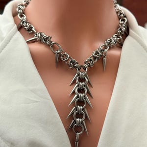 Könnte beinhalten: Eine silberfarbene Halskette mit konisch geformten Spitzen. Die Halskette besteht aus miteinander verbundenen Metallringen und wird auf einer Schaufensterpuppe präsentiert. Die Spitzen sind gleichmäßig verteilt und zeigen nach außen.