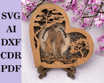 May include: A wooden heart-shaped cutout with a couple dancing inside. The cutout features two trees, a moon, and stars. The couple's names, "Jennifer & Joseph", are written on the moon. The cutout is on a wooden stand.