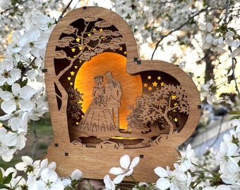 Könnte beinhalten: Herzförmige Hochzeitsdekoration aus Holz mit einer Silhouette von Braut und Bräutigam. Das Design zeigt einen Baum, Sterne und ein warmes, leuchtendes Licht. Die Dekoration ist von weißen Blüten umgeben.