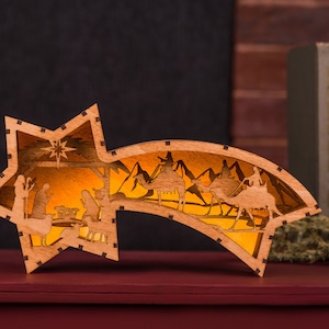 May include: A wooden Christmas star ornament with a cutout design depicting the Nativity scene and the three wise men following a star. The star is illuminated from within, creating a warm glow.