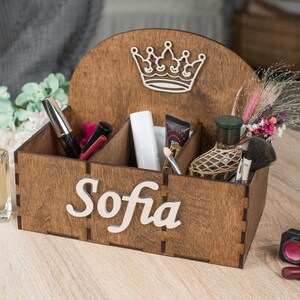 K&ouml;nnte beinhalten: Holzorganizer mit vier F&auml;chern, einem Kronendesign und dem Namen "Sofia" auf der Vorderseite graviert. Der Organizer ist mit verschiedenen Gegenst&auml;nden gef&uuml;llt, darunter Parf&uuml;m, Make-up-Pinsel und Kosmetik.
