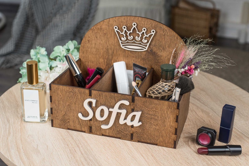 K&ouml;nnte beinhalten: Holzorganizer mit vier F&auml;chern, einem Kronendesign und dem Namen "Sofia" auf der Vorderseite graviert. Der Organizer ist mit verschiedenen Gegenst&auml;nden gef&uuml;llt, darunter Parf&uuml;m, Make-up-Pinsel und Kosmetik.