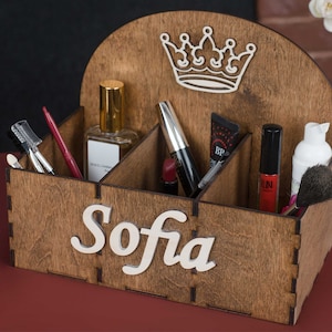 K&ouml;nnte beinhalten: Holzorganizer mit sechs F&auml;chern f&uuml;r Make-up und Kosmetik. Der Organizer ist braun und hat oben ein Kronendesign. Der Name "Sofia" steht auf der Vorderseite des Organizers.