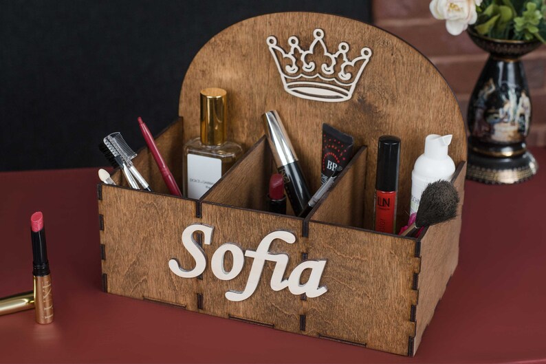 K&ouml;nnte beinhalten: Holzorganizer mit sechs F&auml;chern f&uuml;r Make-up und Kosmetik. Der Organizer ist braun und hat oben ein Kronendesign. Der Name "Sofia" steht auf der Vorderseite des Organizers.