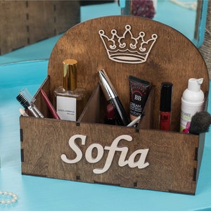 K&ouml;nnte beinhalten: Ein h&ouml;lzerner Make-up-Organizer mit vier F&auml;chern. Der Organizer hat ein Kronendesign auf der Oberseite und den Namen "Sofia" auf der Vorderseite. Der Organizer ist mit verschiedenen Make-up-Artikeln gef&uuml;llt, darunter ein Parf&uuml;mflakon, Mascara, Lippenstift und eine BB-Creme.