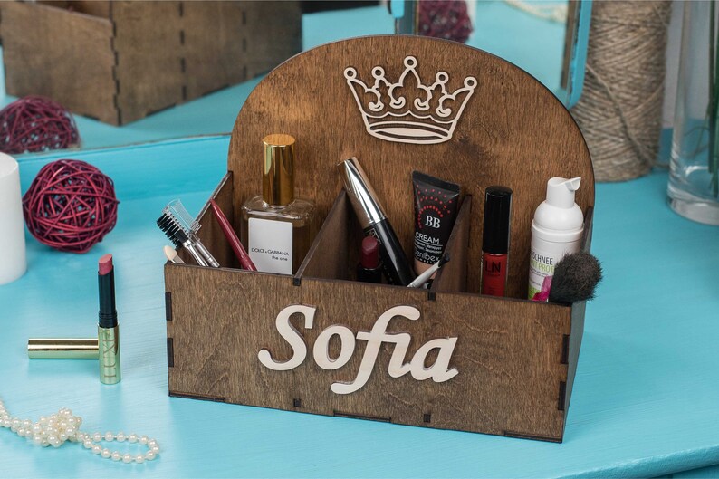 K&ouml;nnte beinhalten: Ein h&ouml;lzerner Make-up-Organizer mit vier F&auml;chern. Der Organizer hat ein Kronendesign auf der Oberseite und den Namen "Sofia" auf der Vorderseite. Der Organizer ist mit verschiedenen Make-up-Artikeln gef&uuml;llt, darunter ein Parf&uuml;mflakon, Mascara, Lippenstift und eine BB-Creme.