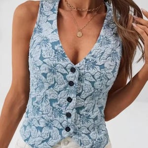 Könnte beinhalten: Eine ärmellose, hellblaue Weste mit floralem Muster und V-Ausschnitt. Die Weste hat eine Knopfleiste mit dunkelblauen Knöpfen. Das florale Design ist in einem helleren Blauton gehalten und wirkt strukturiert.