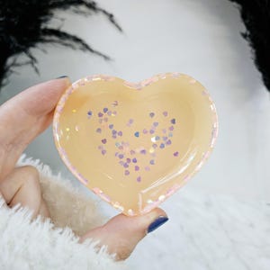 May include: A heart-shaped dish made of clear resin with a light peach colour and iridescent pink and purple glitter hearts.