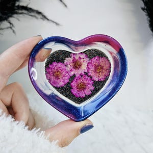 May include: A heart-shaped resin dish with a blue, pink and white swirl design. The dish contains three pressed pink flowers on a black glitter background.
