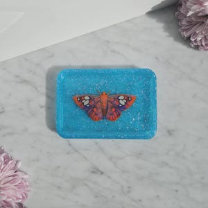 May include: A rectangular, turquoise resin tray with a glittery finish. The tray features a detailed butterfly design in shades of orange, purple, and white. The butterfly is centered on the tray, creating a decorative accent.