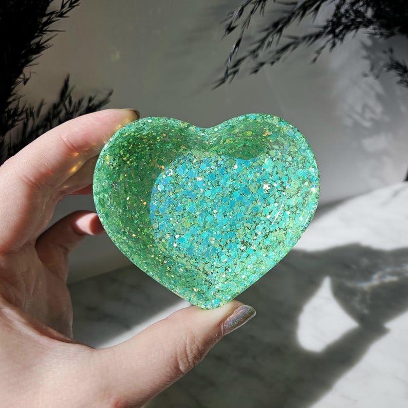 May include: A heart-shaped dish made of clear resin with green and blue glitter embedded throughout. The dish is ideal for holding small items such as jewellery or trinkets.