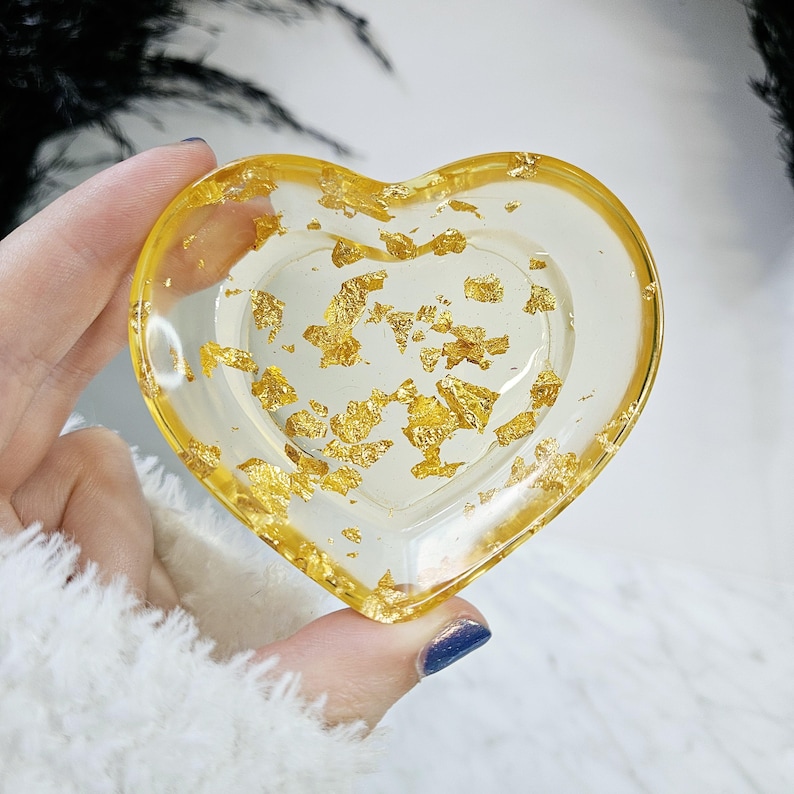 May include: A heart-shaped resin tray with gold leaf flakes embedded inside. The tray is clear and has a slight yellow tint.