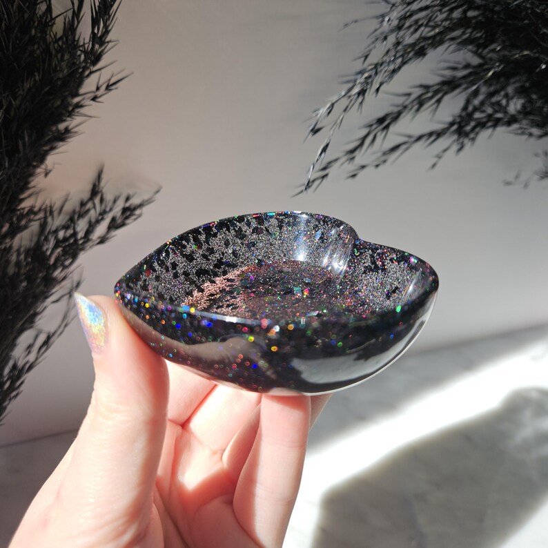 May include: A black heart-shaped dish with iridescent glitter embedded in the resin. The dish is made of resin and has a smooth, glossy finish.