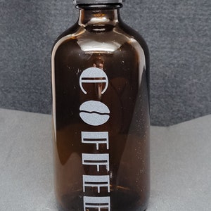 Op de afbeelding: Een bruine glazen fles met een zwarte dop. De fles heeft het woord "COFFEE" verticaal in het wit gedrukt, samen met een koffieboongrafiek. De fles is ontworpen voor koffieopslag.