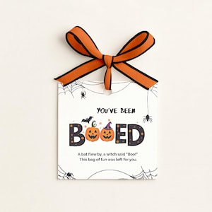 May include: A white Halloween gift tag with an orange and black ribbon. The tag says "YOU'VE BEEN BOOED" with pumpkin and bat graphics. Text at the bottom reads "A bat flew by, a witch said 'Boo!' This bag of fun was left for you."