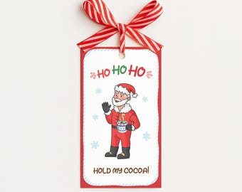 May include: A red gift tag with a cartoon Santa Claus holding a mug of cocoa. The tag has the text "HO HO HO" and "HOLD MY COCOA!" with a red and white striped ribbon at the top.