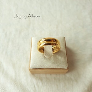 May include: A gold ring with a double band design, displayed on a white cushion in a wooden box. The ring is made of a smooth, shiny metal.