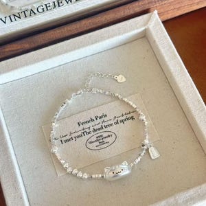 May include: A silver bracelet with a cat charm and small beads. The bracelet is displayed in a white box with the text "Paris VINTAGE JEWELRY COLLECTOR". A tag with the letter "L" hangs from the bracelet.
