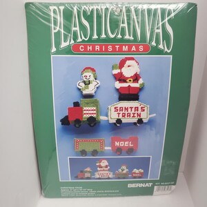 May include: A plastic canvas Christmas train kit with Santa, a snowman, and a train car that says "Santa's Train". The kit includes yarn, needles, charts, and instructions. Bernat Kit 95-0527-00.