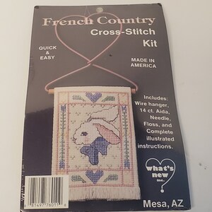 Puede incluir: Un kit de punto de cruz con un conejo blanco con detalles azules y rosas. El kit incluye un colgador de alambre, tela Aida de 14 puntos, aguja, hilo y instrucciones ilustradas completas. El kit está hecho en Estados Unidos y es rápido y fácil de completar. El empaque presenta el texto "French Country Cross-Stitch Kit" y "What's New Inc."