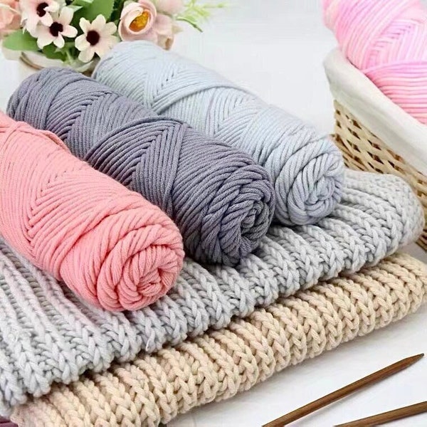 May include: Four balls of yarn in pink, gray, and light gray colors. The yarn is arranged on top of a knitted fabric in beige, gray, and light gray colors.