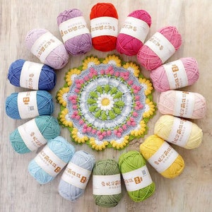 Puede incluir: Una colecci&oacute;n de ovillos de lana coloridos en varios tonos, incluyendo morado, rojo, rosa, azul, verde y amarillo, dispuestos alrededor de un tapete de ganchillo. El tapete presenta un dise&ntilde;o floral con detalles rosas y verdes. Los ovillos de lana tienen etiquetas con caracteres chinos.