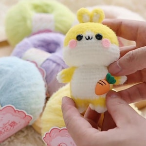 Puede incluir: Un conejito de crochet amarillo y blanco sosteniendo una peque&ntilde;a zanahoria naranja. El conejito tiene ojos negros y mejillas rosadas. El conejito est&aacute; rodeado de ovillos de lana de varios colores.